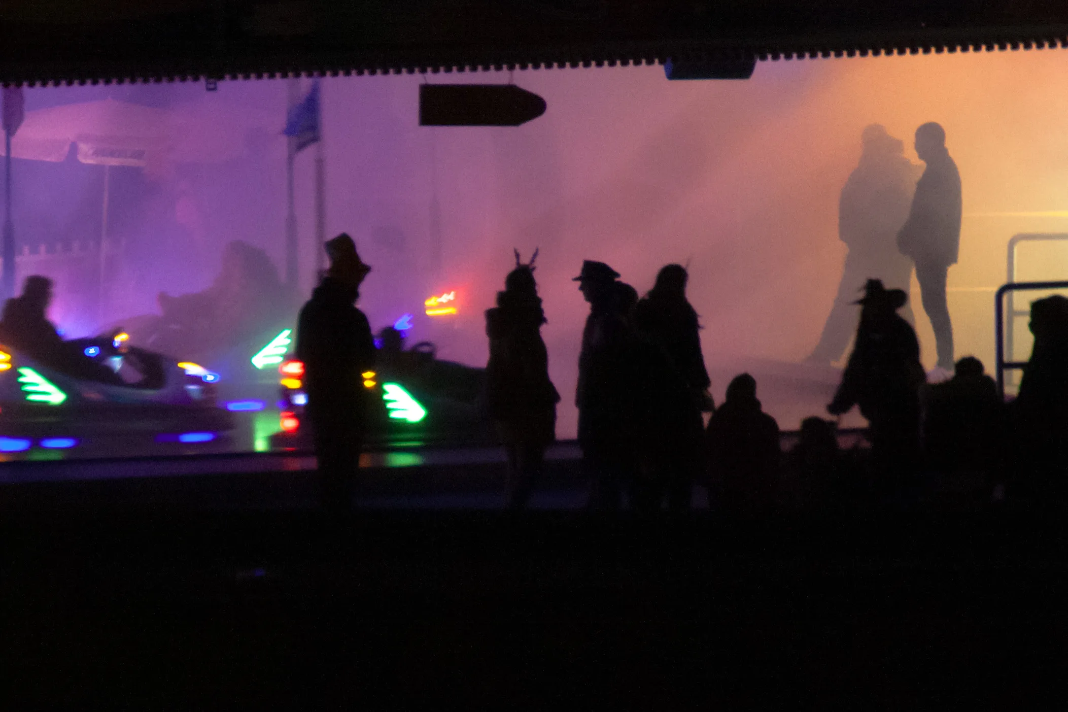 A nighttime telephoto of a crowd gathered around a bumper car ride at a small-time amusement park. The crowd is unlit, and seen only as black silhouettes against the backdrop of the bumper car ride. Smoke is covering part of the ride, catching the light from the park and the bumper cars.