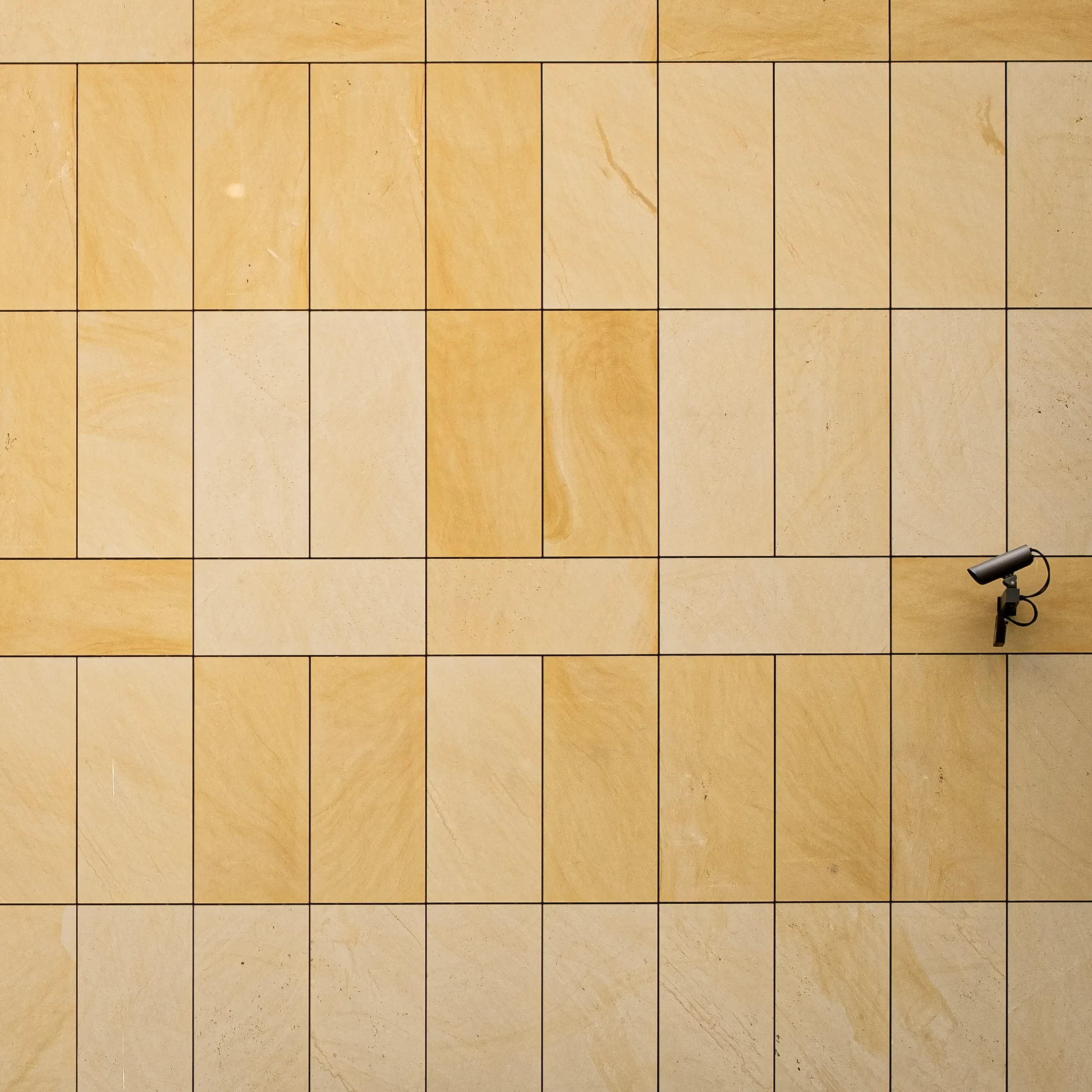A photo of the exterior wall of a building, with a security camera looking along the wall. The wall is covered by a grid-like arrangement of large rectangular tiles, each with a slightly different shade of yellow and natural texture, creating an interesting geometrical texture.