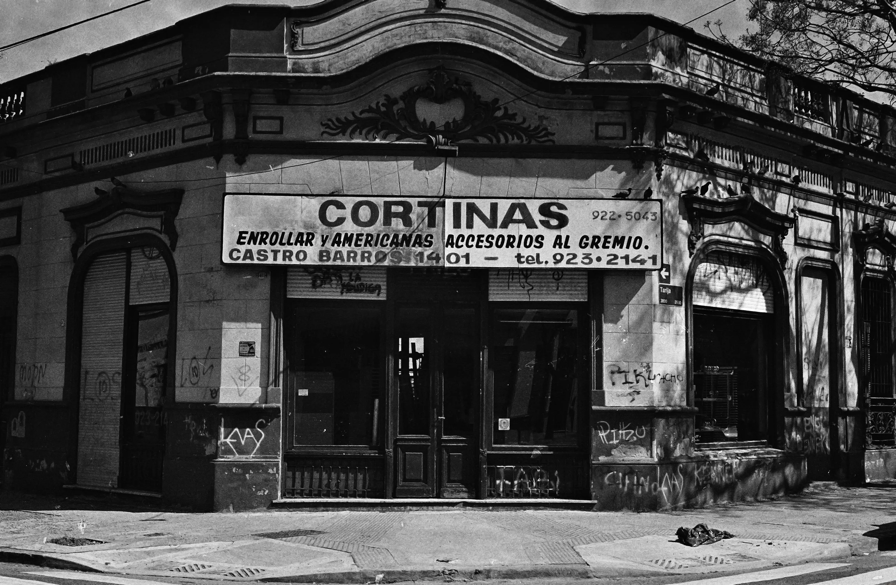 A black and white photo of a traditional street corner in Buenos Aires. There's a store directly in the corner, but it's unclear if it's still in business, and the walls are covered in grafitti tags.