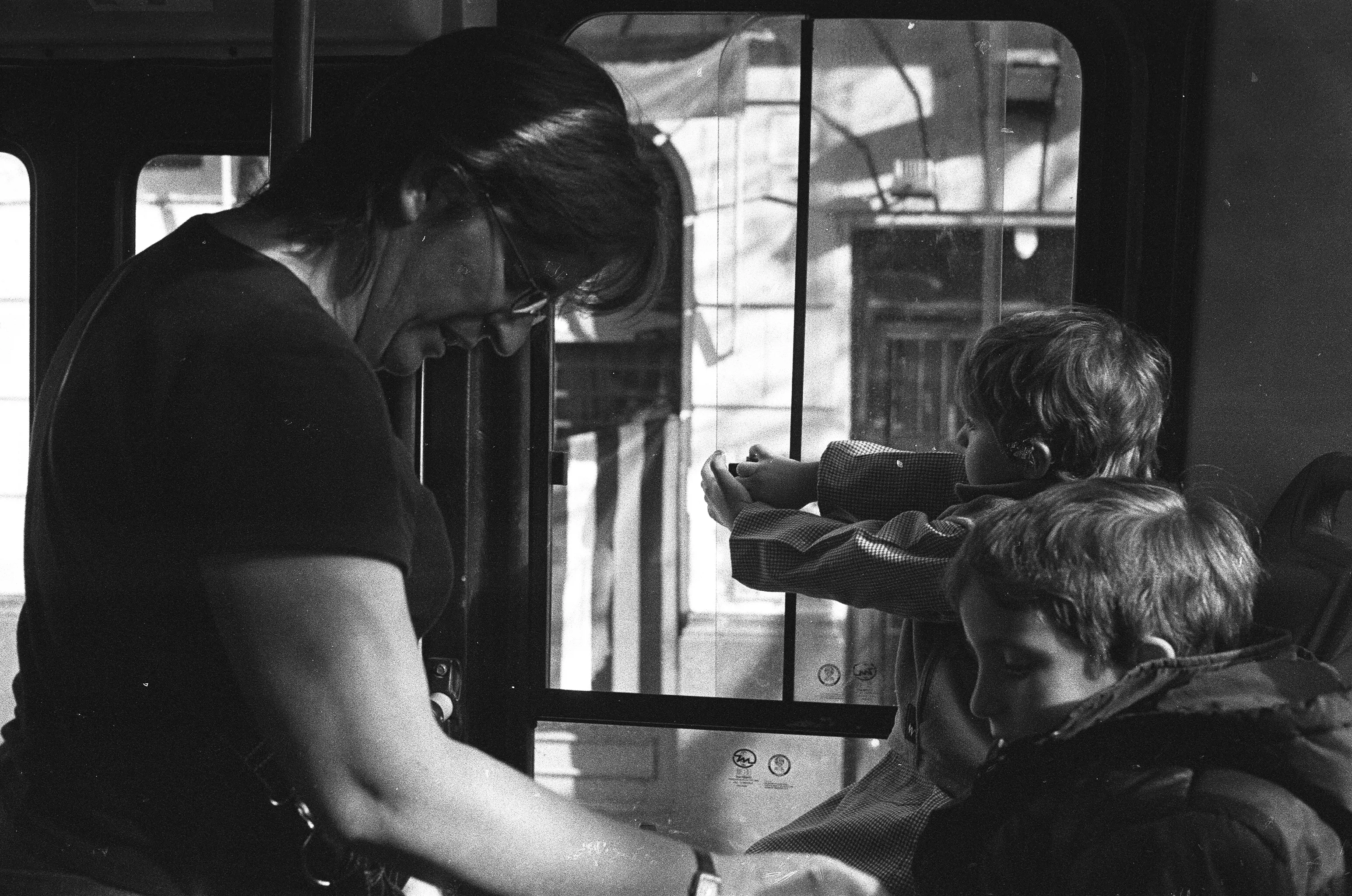 A black and white photo of the inside of a bus. Two kids are sitting down, with a lady tending to one of them while the second kid is sliding the bus window open.