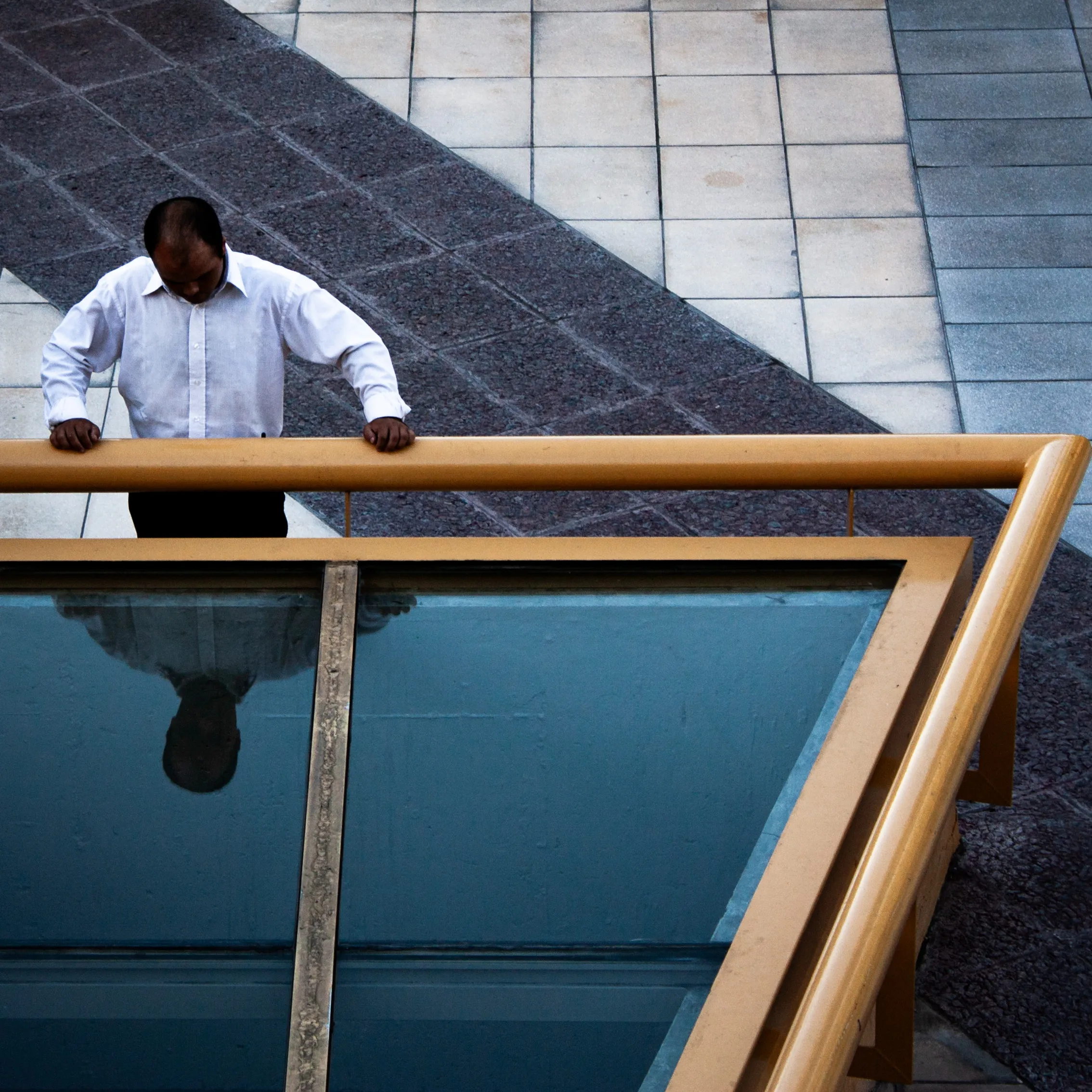A man in a white shirt is looking down a glass ceiling. He's resting against a bright yellow handrail that creates a sharp angle across the frame. We can see his reflection in the glass looking back at him.