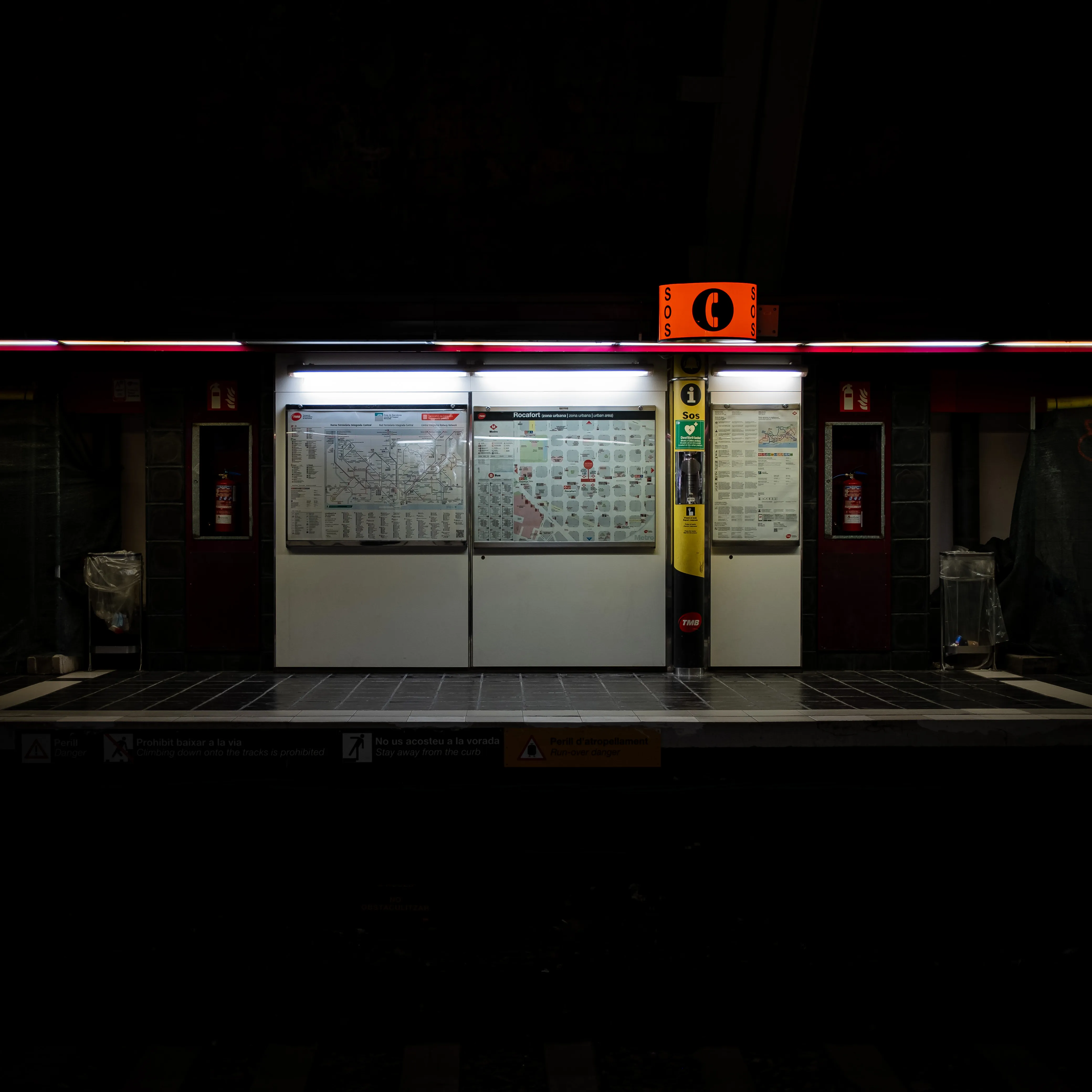 An information stall inside a Barcelona subway station. There's a bright red SOS sign, a yellow telephone, and two well-lit maps.
