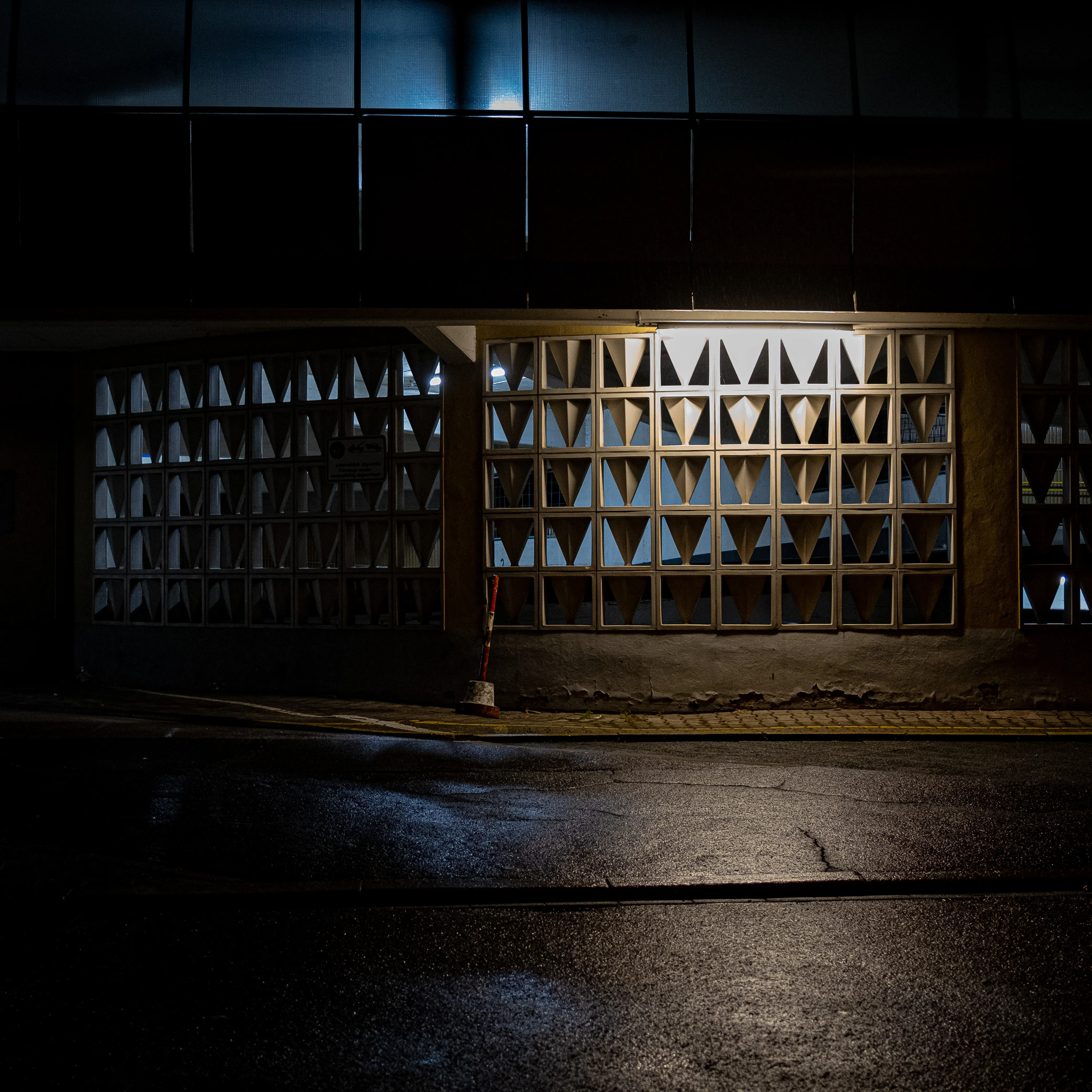 The street outside of a parking garage, during the night. A dim light shines from the parking garage wall, higlighting its geometric patterns. The pavement is wet from the rain.